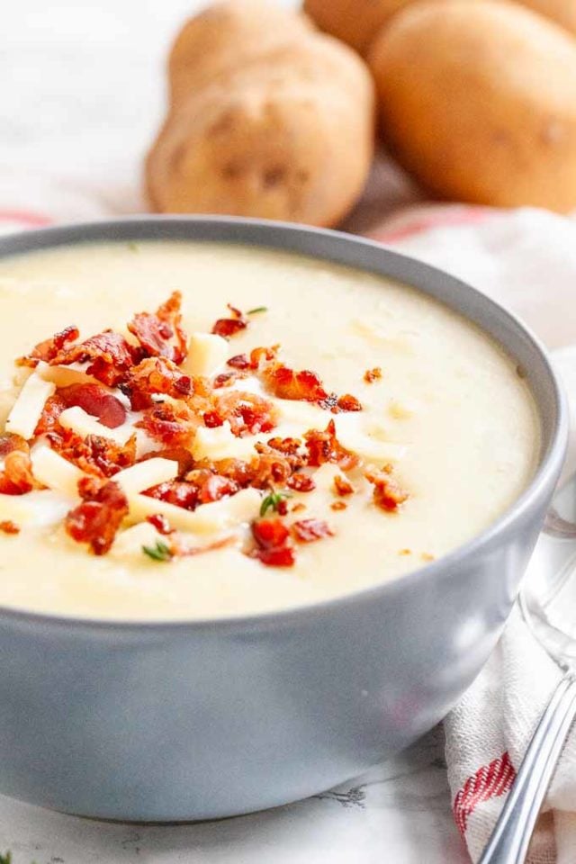 A grey bowl of potato soup topped with bacon crumbs, cheese stripes and thyme. Potatoes in background.