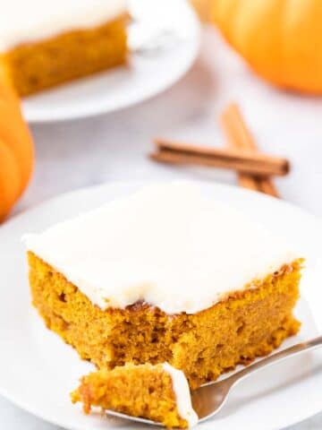 A piece of pumpkin cake topped with frosting and a pecan nut on a white plate. A fork has taken out a piece and is lying next to it. There are 2 pieces of cinnamon and two small pumpkins in the background.