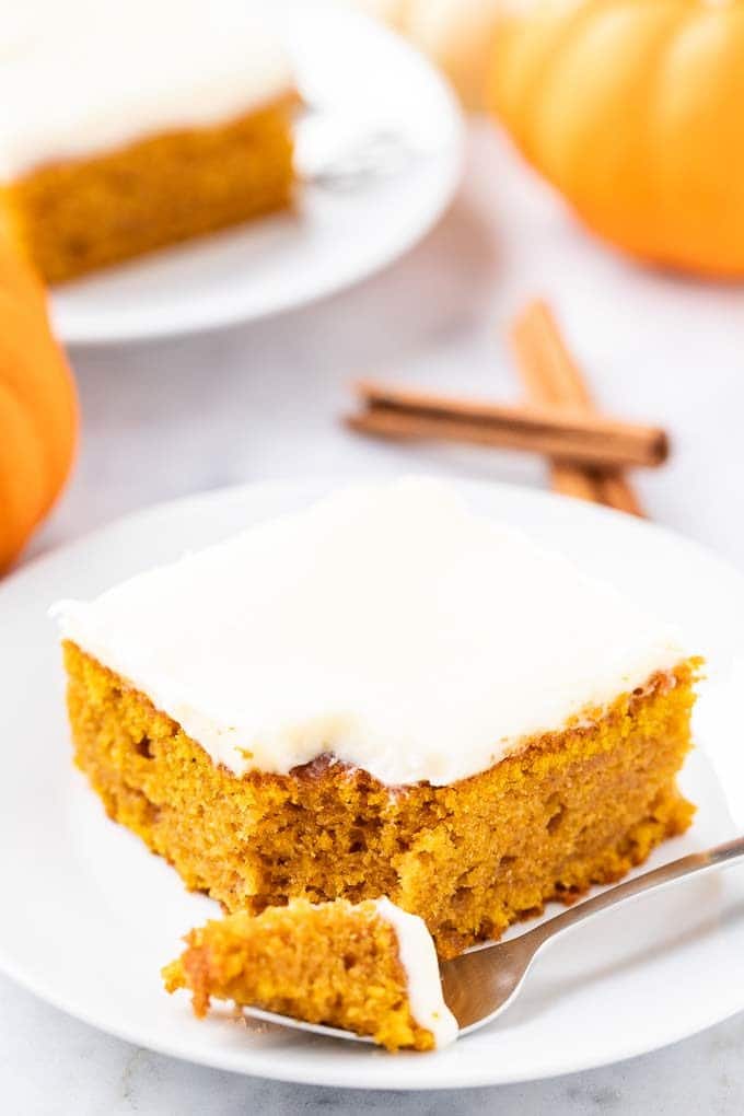 A piece of pumpkin cake topped with frosting and a pecan nut on a white plate. A fork has taken out a piece and is lying next to it. There are 2 pieces of cinnamon and two small pumpkins in the background.