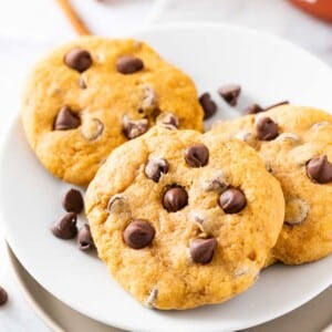 Three chocolate chip cookies on a white plate, garnished with chocolate chips on a marble cutting board.