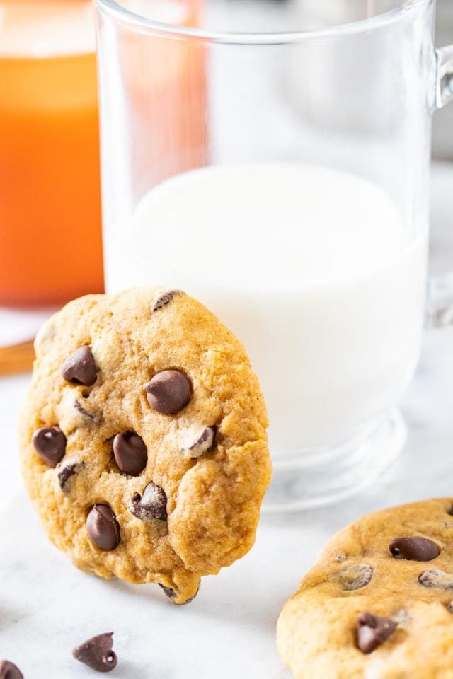A chocolate chip cookie leaning against a glass of milk, with a cookie lying next to it.
