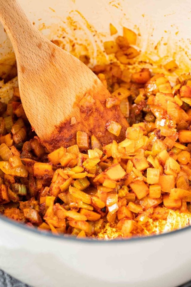 A dutch oven with diced onions, carrots, celery and paprika powder being sauteed with a wooden spatula in it.
