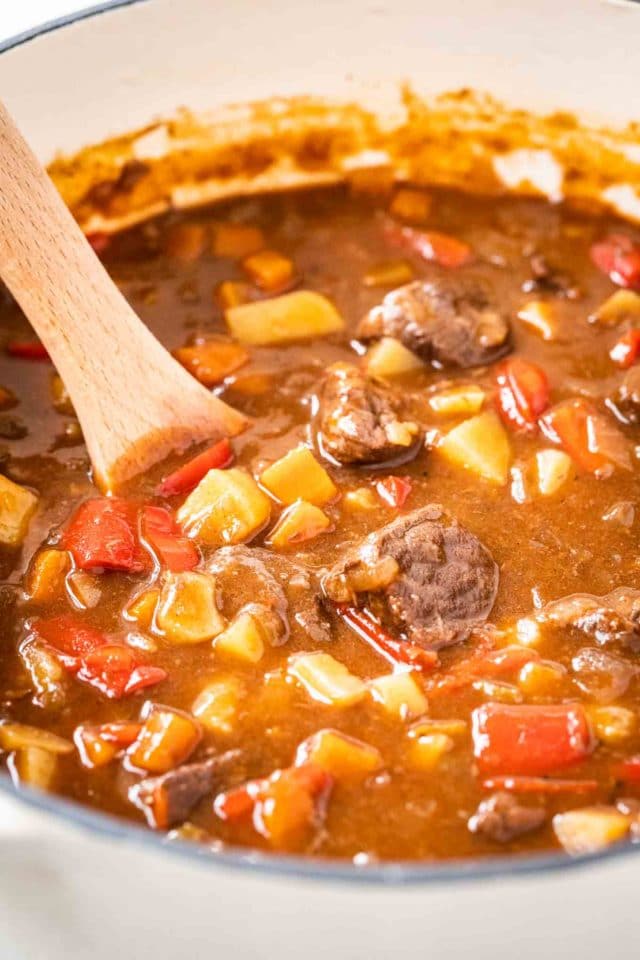 A dutch oven with Goulash, containing red peppers, beef and potatoes with a wooden spoon in it.