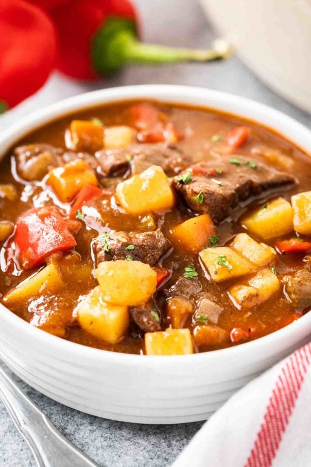 A white bowl of goulash, containing red peppers, beef and potatoes, topped with parsley. There\'s a white dish towel with red stripes and some red peppers next to it.