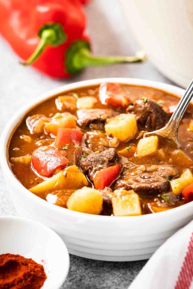 A white bowl of goulash, containing red peppers, beef and potatoes, topped with parsley and a spoon in it. There\'s a white dish towel with red stripes and some red peppers next to it.