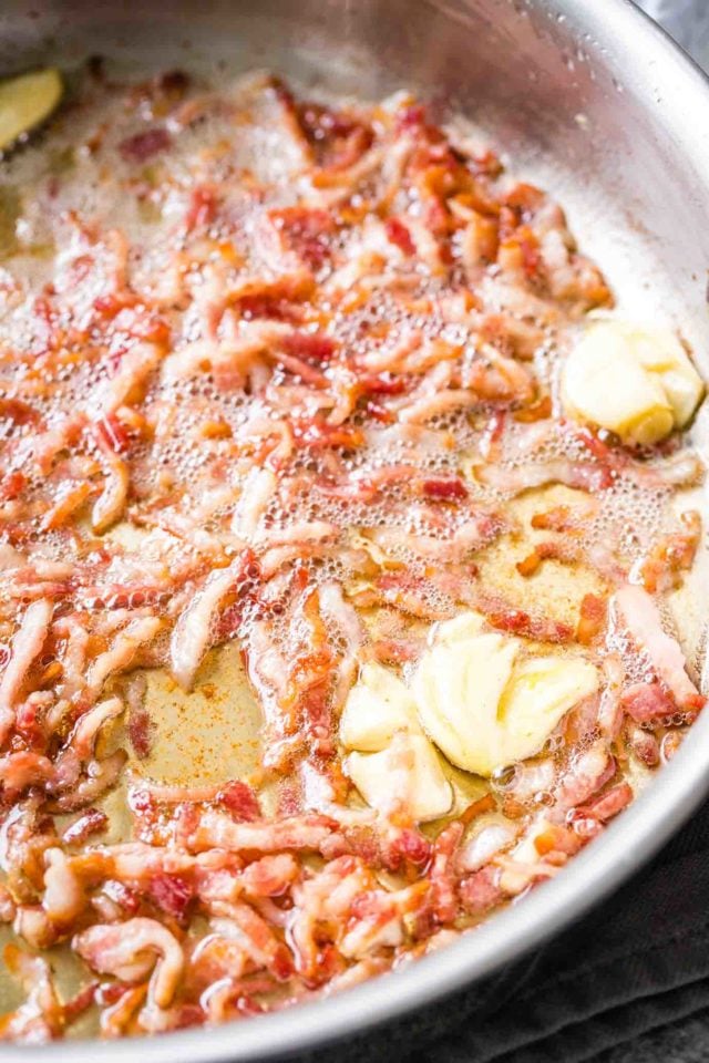 Bacon, cut into small pieces, being fried in a stainless tell pan, with some crushed and peeled garlic cloves.
