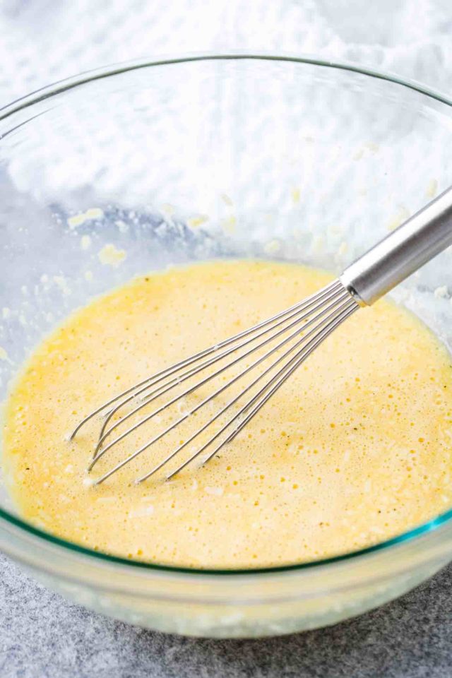 A glass bowl with a stainless tell whisk containing a mixture of egg and grated parmigiano