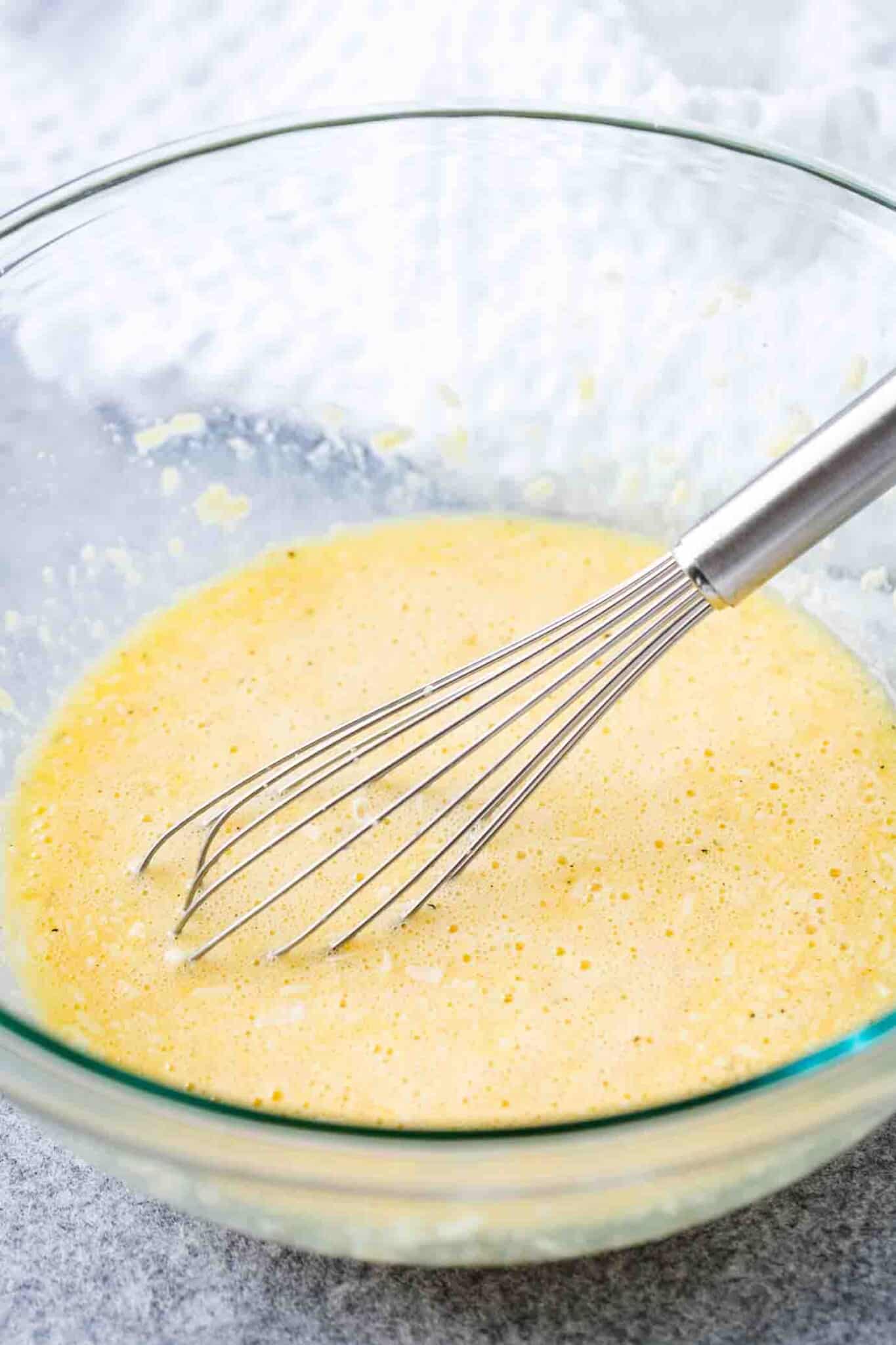 A glass bowl with a stainless tell whisk containing a mixture of egg and grated parmigiano.