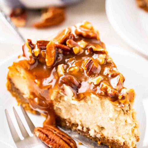 A slice of cheesecake topped with pecan nuts and caramel sauce on a small white plate with a fork.