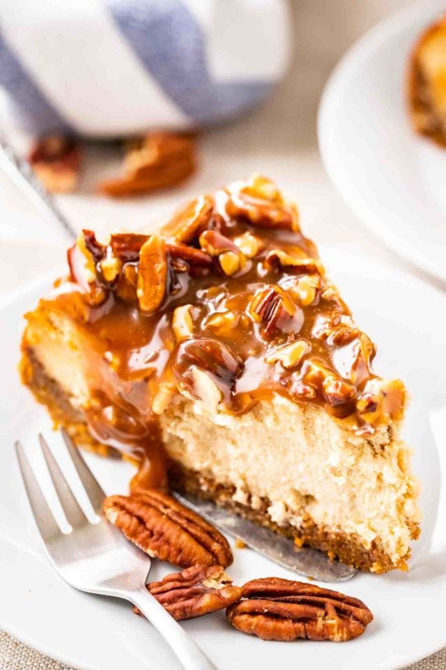 A slice of cheesecake topped with pecan nuts and caramel sauce on a small white plate with a fork. 