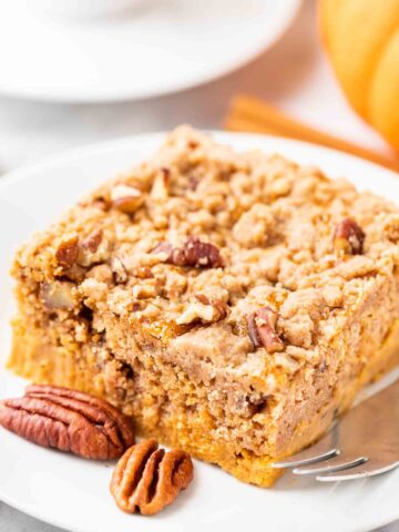 A piece of pumpkin cake with pecans on a white plate with a fork, garnished with whole pecans, cinnamon sticks and pumpkin.