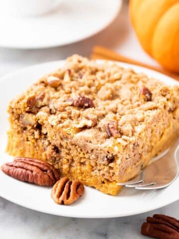 A piece of pumpkin cake with pecans on a white plate with a fork, garnished with whole pecans, cinnamon sticks and pumpkin. There\'s a cup of espresso in the background