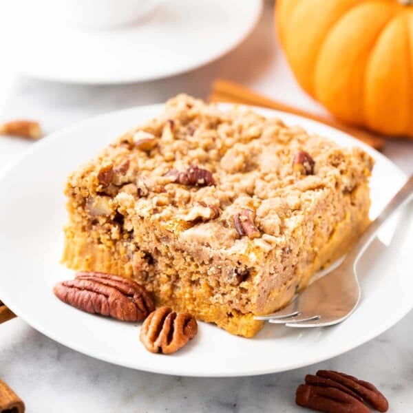 A piece of pumpkin cake with pecans on a white plate with a fork, garnished with whole pecans, cinnamon sticks and pumpkin. There\'s a cup of espresso in the background