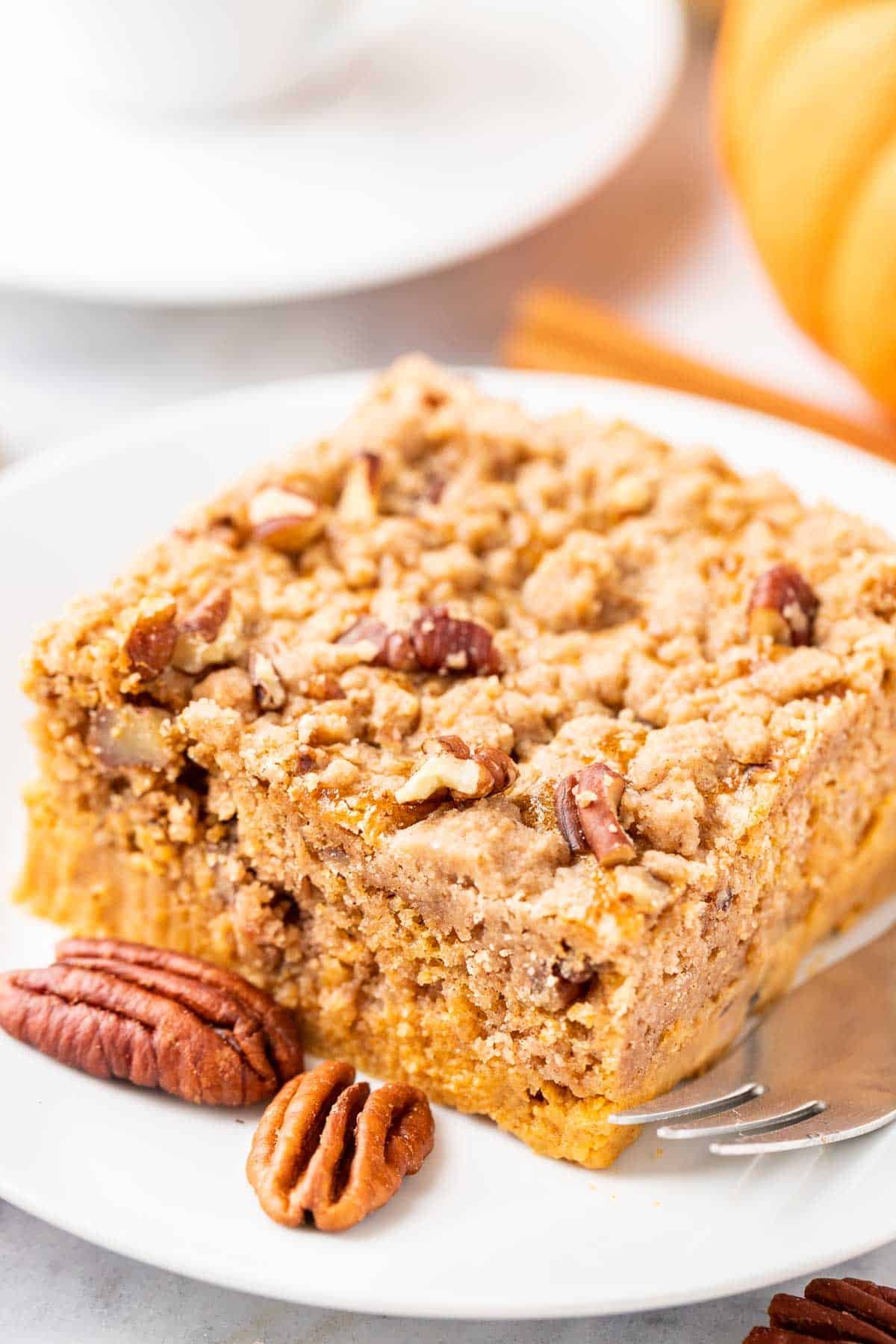A piece of pumpkin cake with pecans on a white plate with a fork, garnished with whole pecans, cinnamon sticks and pumpkin.