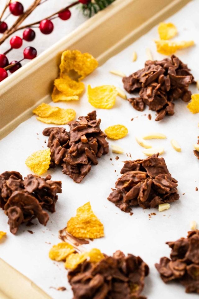 chocolate covered clusters of corn flakes and almonds on parchment paper in a baking pan, garnished with corn flakes, almonds and cranberries