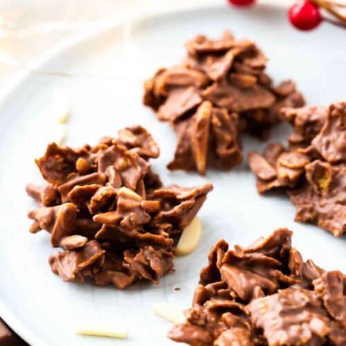 chocolate covered clusters of corn flakes and almonds on a grey plate, garnished with almonds and cranberries