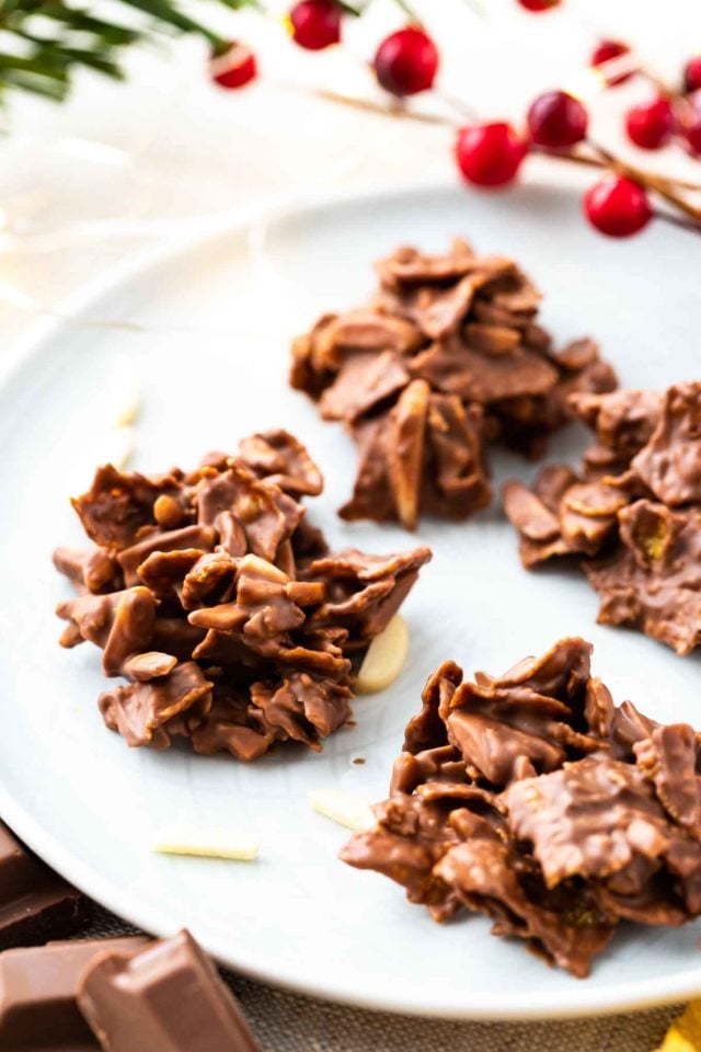 chocolate covered clusters of corn flakes and almonds on a grey plate, garnished with almonds and cranberries