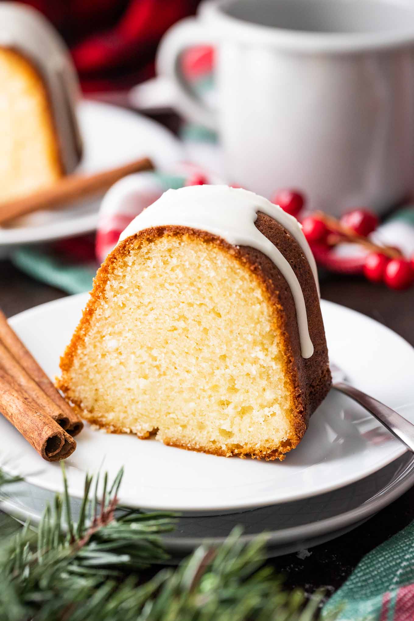 A piece of bundt cake with white glazing on top on a white plate with a fork and two sticks of cinnamon. There are cranberries, a cup and another plate in the background.