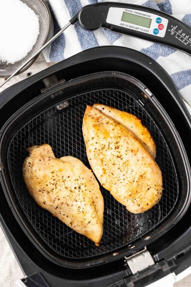 Two fried chicken breasts sitting in an air fryer basket on a white and blue dish towel with a dark bowl with salt and a thermometer next to it.