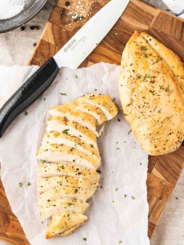 Two fried chicken breasts on a dark wood cutting board and parchment paper with a ceramic knife next to it. One chicken breast is sliced.