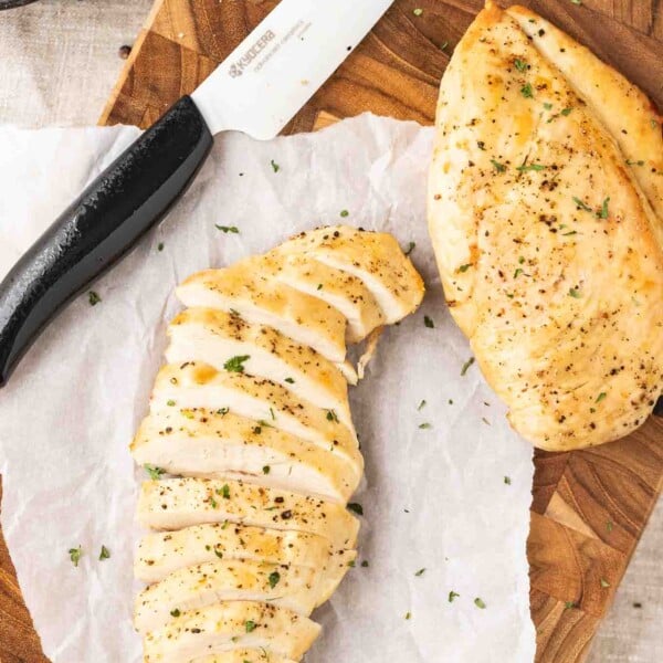 Two fried chicken breasts on a dark wood cutting board and parchment paper with a ceramic knife next to it. One chicken breast is sliced.