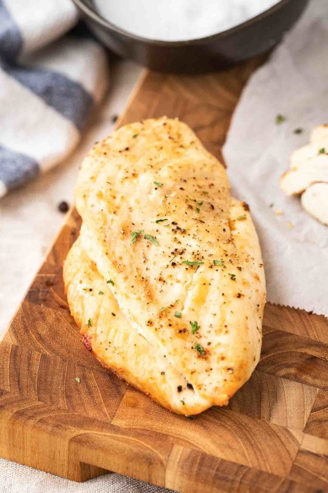 A grilled chicken breast with herbs sitting on a dark wood cutting board next to some sliced chicken breast and a white and blue dish towl.