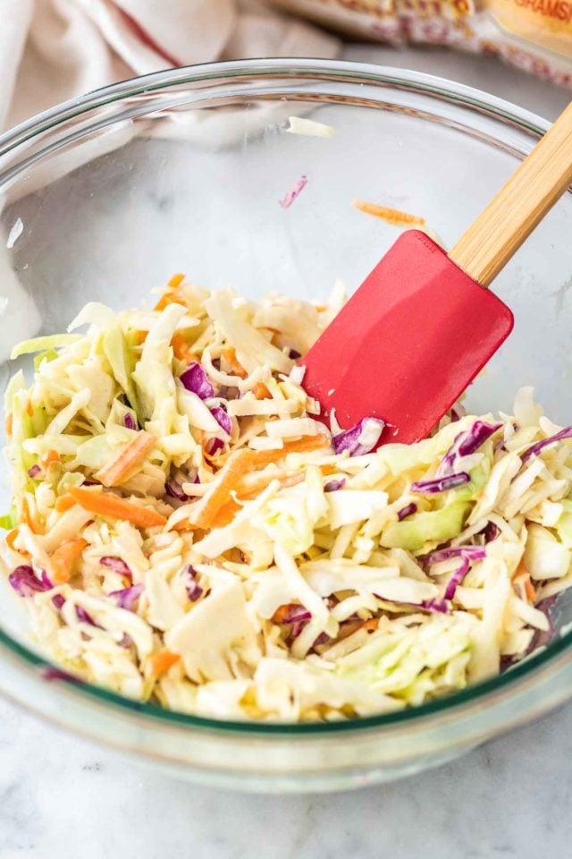 A glass bowl of coleslaw with a red spatula with a wooden handle in it.