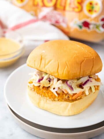 A burger with a breaded fried chicken patty and coleslaw on a white plate next to a glass bowl with dip, a white and red towel and a bag of burger buns