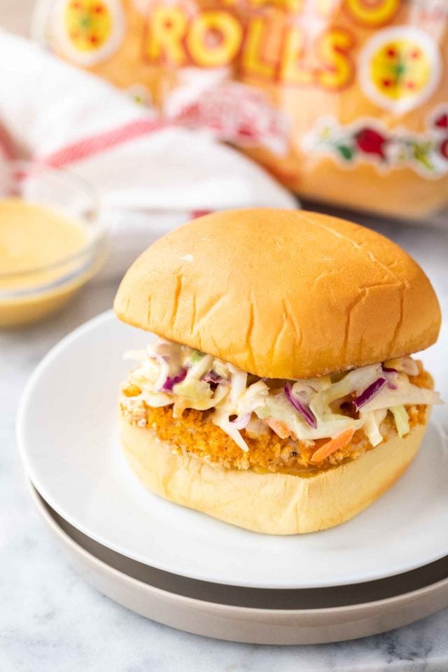 A burger with a breaded fried chicken patty and coleslaw on a white plate next to a glass bowl with dip, a white and red towel and a bag of burger buns