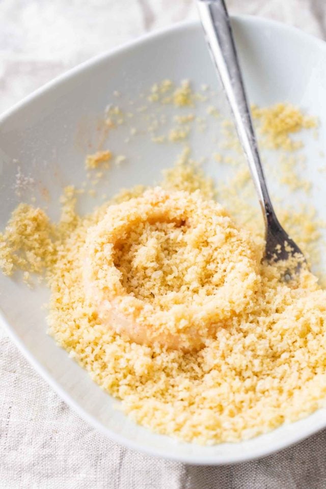 an onion ring in some panko crumbs in a white plate with a fork in it.