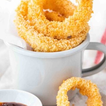 Fried onion rings in a grey canteen cup with parchment paper. There's a small white porcelain bowl with barbecue sauce next to it and a single onion ring leaning against it.
