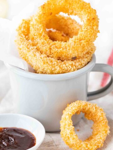 Fried onion rings in a grey canteen cup with parchment paper. There's a small white porcelain bowl with barbecue sauce next to it and a single onion ring leaning against it.