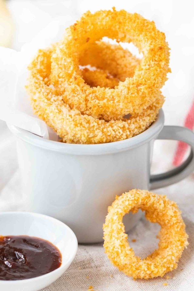Fried onion rings in a grey canteen cup with parchment paper. There\'s a small white porcelain bowl with barbecue sauce next to it and a single onion ring leaning against it.