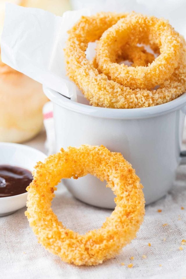 A doughnut is next to a cup of coffee, with Onion ringFried onion rings in a grey canteen cup with parchment paper. There\'s a small white porcelain bowl with barbecue sauce next to it, a whole raw onion in the background and a single onion ring leaning against it.