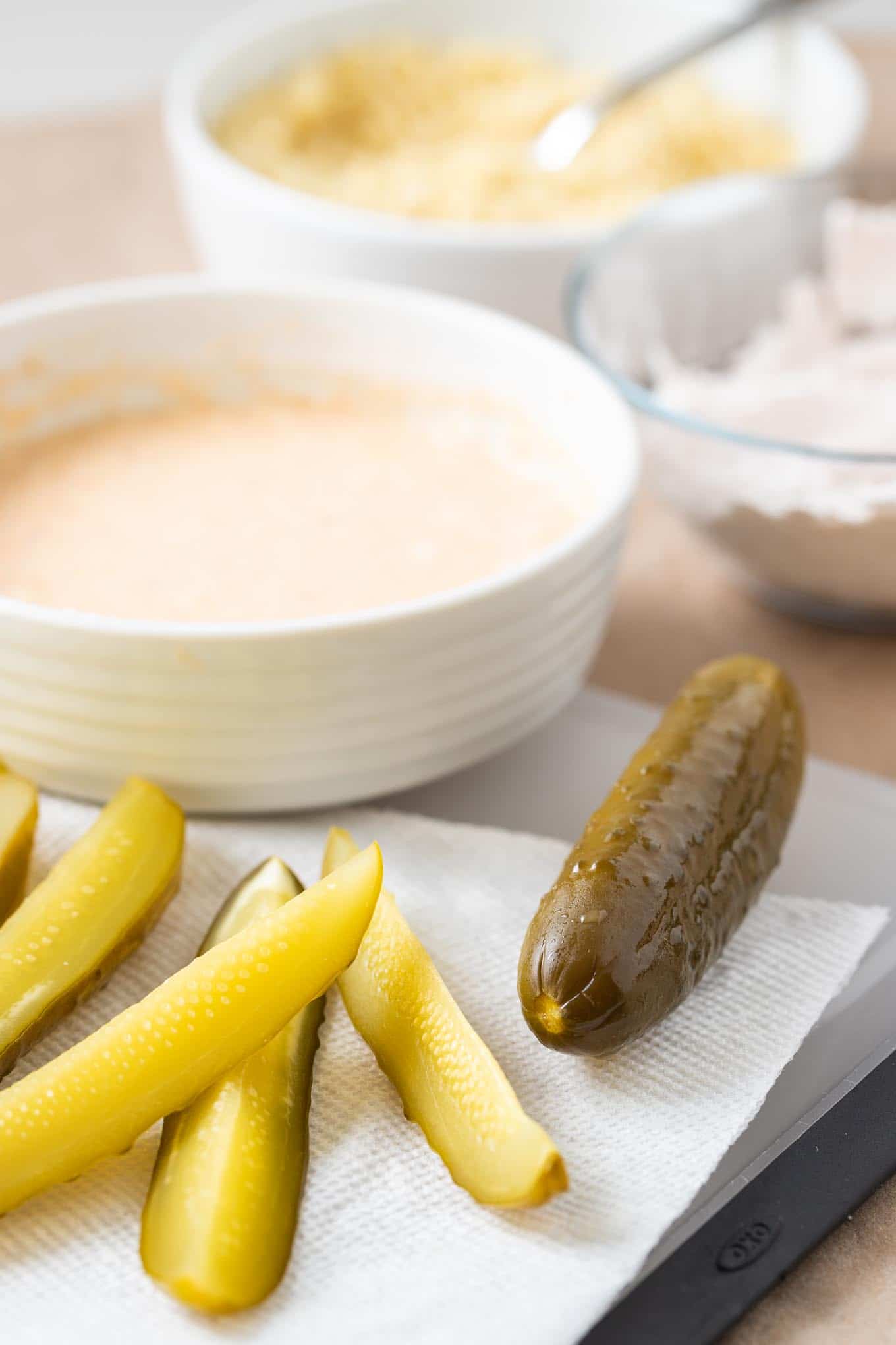 Dill pickle wedges, with a whole dill pickle on a cutting board with a kitchen towel. There\'s a white bowl with batter and a glass bowl with flour in the background.