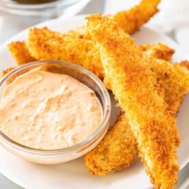 A heap of air fryer dill pickles on a white plate with a small glass bowl with a dip in it.