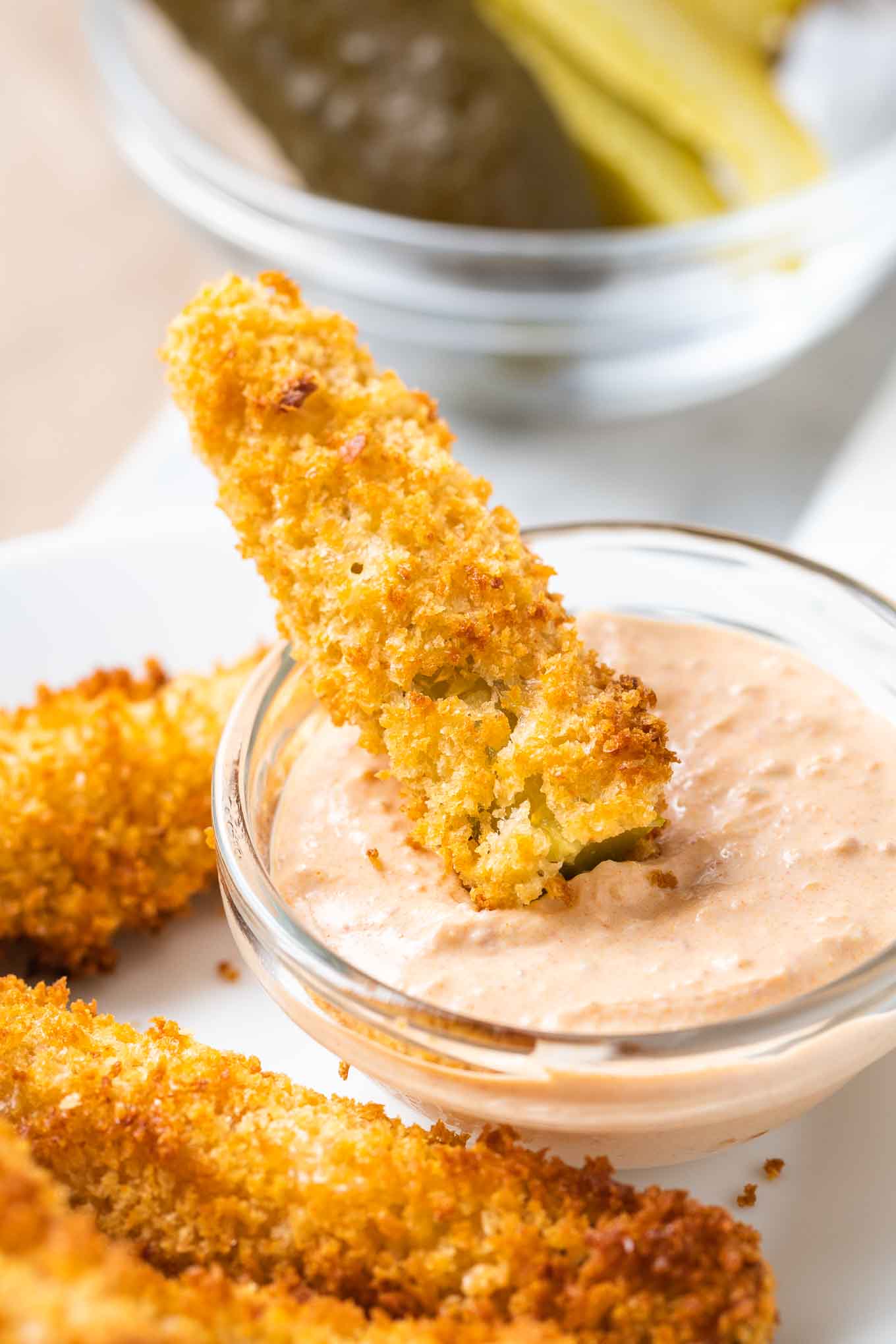 A fried dill pickle is dipped into sauce in a small glass bowl, with more fried pickles next to it on a white plate.