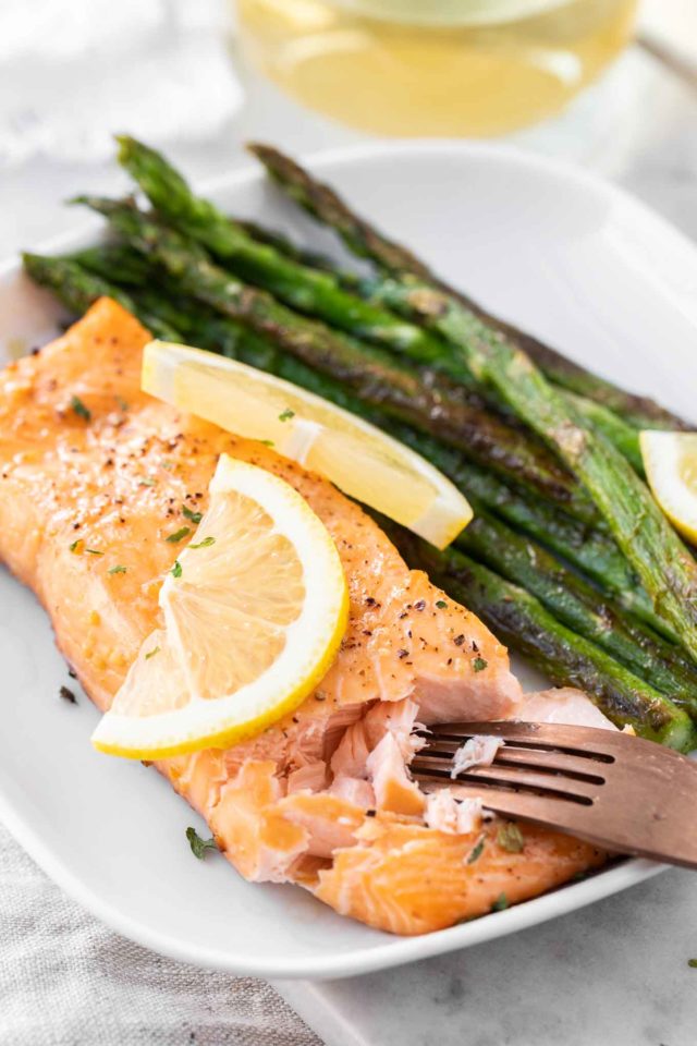 A white plate with a piece of cooked salmon next to roasted green asparagus, garnished with lemon slices. A bronze fork is flaking the salmon.