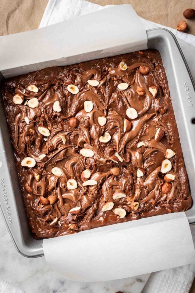 Baking tray lined with parchment paper containing Nutella brownies topped with hazelnuts
