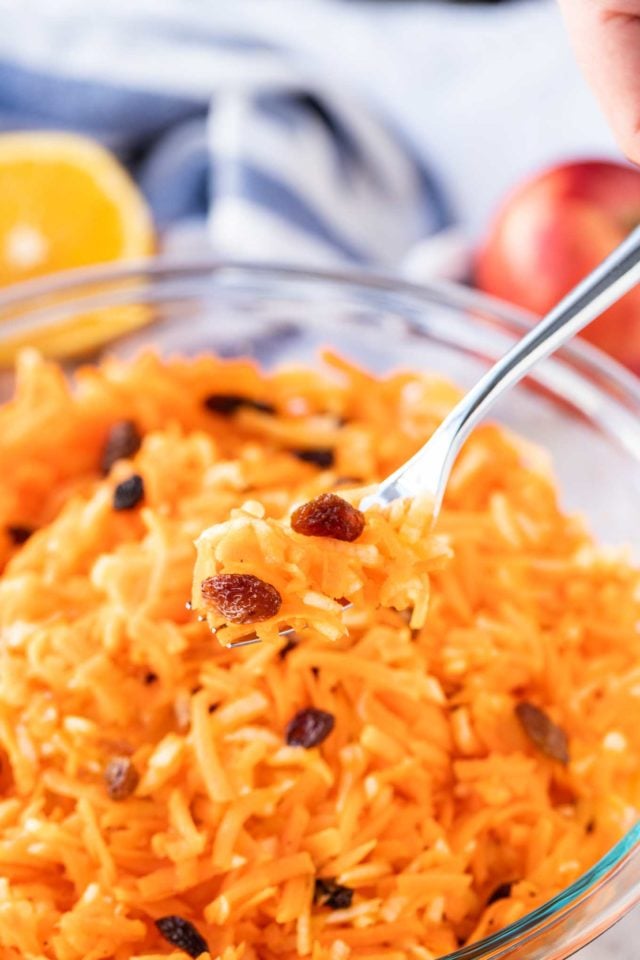 Carrot salad with raisins in a glass bowl, with a portion of it on a fork hovering above it. Half and orange and an apple in the background