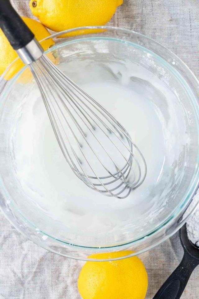 Lemon glaze in a glass bowl with a whisk in it and a lemon and a bowl of powdered sugar next to it