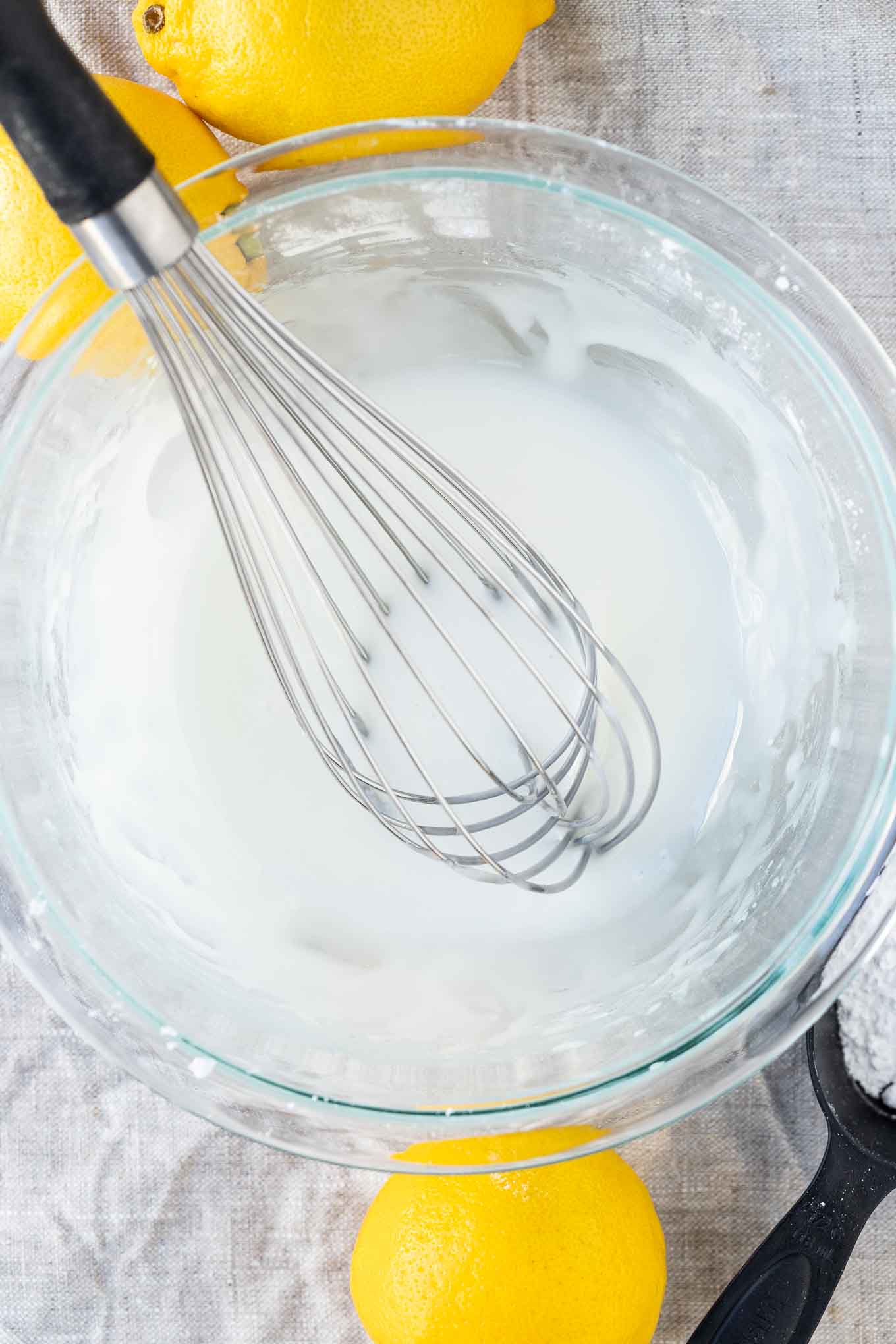 Lemon glaze in a glass bowl with a whisk in it and a lemon and a bowl of powdered sugar next to it.
