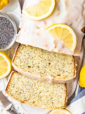 top-down view of lemon poppy seed cake with two slices cut off on a white ceramic cutting board, garnished with lemons next to a glass bowl with poppy seeds