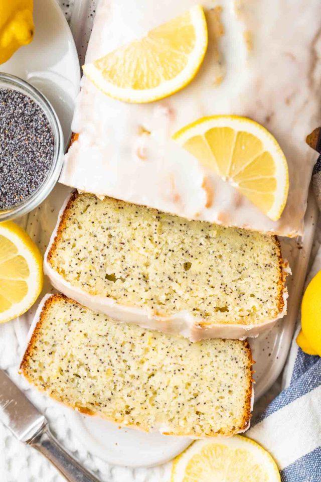 top-down view of lemon poppy seed cake with two slices cut off on a white ceramic cutting board, garnished with lemons next to a glass bowl with poppy seeds