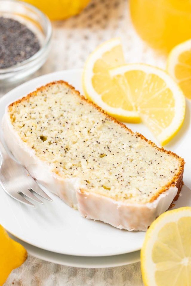 a slice of lemon poppy seed cake on a white plate with a fork, garnished with lemon slices and a glass bowl with poppy seeds in the background.