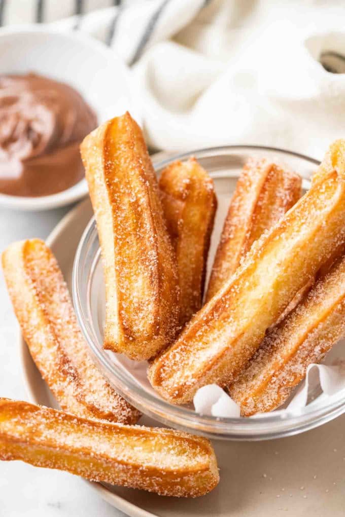 sugared churros in a glass bowl with parchment with a small white bowl of nutella in the background