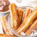 sugared churros in a glass bowl with parchment with a small white bowl of nutella in the background