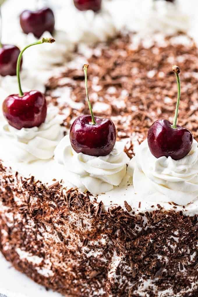 A close-up of a Black Forest Cake. The cake has chocolate shavings on the side and swirls of whipped cream topped with whole cherries on top
