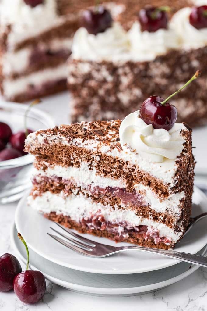 A piece of black forest cake on a white plate next to a cake fork. In the background is a small glass bowl filled with cherries and a cake