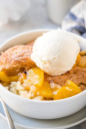 Close-up of a bowl of peach cobbler in a white bowl with a scoop of vanilla ice cream on top